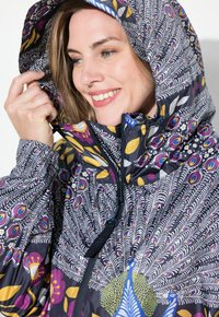 Veste imperméable colorée avec un motif de plumes de paon, dotée d'un col haut, d'une capuche ajustable et d'une fermeture éclair. Tissu doux et léger.