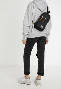 Borsa a tracolla nera con chiusura a zip e dettagli in marrone, indossata sopra una felpa grigia e jeans neri, abbinata a sneakers grigie chiare.