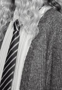 Grå stickad kofta, vit plisserad skjorta med vertikala ränder och ett svart slips med diagonala vita ränder. Håret är långt och vågigt.