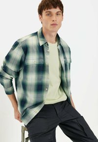 Grøn- og blåternet skjorte med lange ærmer og knaplukning; blødt stof, to brystlommer, båret over en lysegrøn t-shirt.