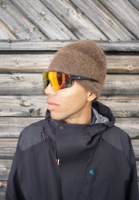 Μαύρο μπουφάν με κουκούλα, καφέ σκουφάκι από υφή τσόχας και γυαλιά ηλίου με γωνιώδη πορτοκαλί απόχρωση. Το φόντο από ξύλινο σχέδιο δημιουργεί αντίθεση.