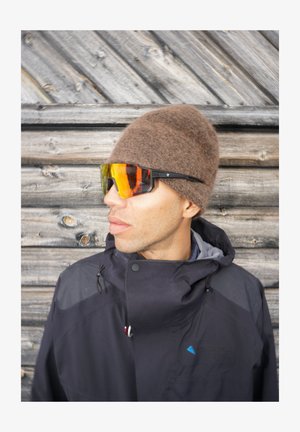 Černá bunda s kapucí, hnědá čepice z plstěné tkaniny s texturou a hranaté sluneční brýle s oranžově tónovanými skly. Pozadí s dřevěným vzorem vytváří kontrast.