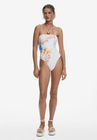 Maillot de bain sans bretelles en blanc, orné d'impressions colorées de coquillages et d'étoiles de mer. Mette en valeur des fines bretelles roses et un pendentif en or.