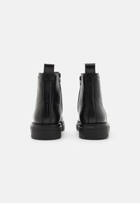 Une paire de bottes noires en cuir à la cheville avec des tirettes à l'arrière et des zips sur les côtés, présentées de dos sur un fond blanc uni.