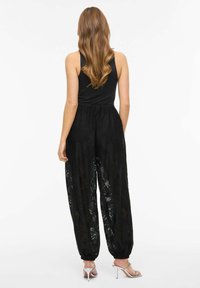 Czarny kombinezon z koronki o luźnym kroju, z marszczoną talią i elastycznymi mankietami. Posiada gorset w stylu tank top oraz przezroczysty kwiatowy wzór. Srebrne szpilki.