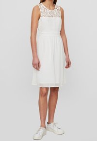 Robe blanche sans manches avec un corsage en dentelle et une taille élastique. La jupe arrive au-dessus du genou. Portée avec des baskets blanches.