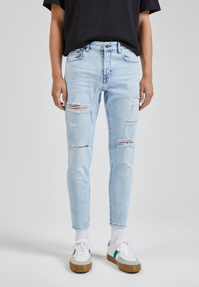 Personne portant un jean skinny déchiré bleu clair, des chaussettes blanches, des baskets blanches avec un détail bleu sarcelle, et un t-shirt noir court au niveau des hanches.