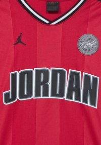 Raudona maikė su juodais akcentais, su juodu Jumpman logotipu ir dideliu „JORDAN“ tekstu. Pagaminta iš tekstūruoto audinio su vertikaliais dryžiais.
