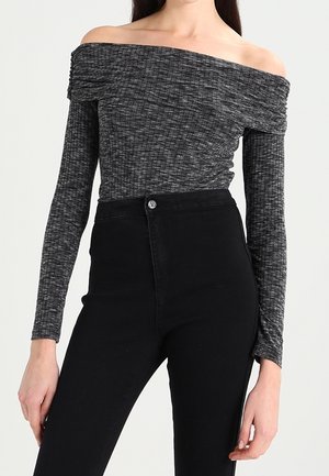 Femme portant un haut gris à manches longues et épaules dénudées et un pantalon noir taille haute sur un fond uni.