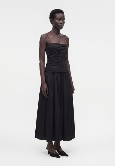 Γυναίκα με μαύρο τοπ με λεπτές τιράντες και ρυτιδωτό ύφασμα, μακριά μαύρη πλισέ φούστα και μαύρα μυτερά παπούτσια με χαμηλό τακούνι, στέκεται μπροστά σε απλό φόντο.
