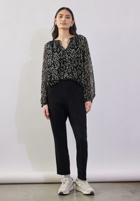 Durchsichtige schwarze Bluse mit einem weißen geometrischen Muster, langen Ärmeln und einem lässigen Schnitt, kombiniert mit schwarzen maßgeschneiderten Hosen und weißen Sneakers.