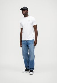 Camiseta branca de mangas curtas com um logótipo subtil no peito, combinada com jeans azuis de pernas direitas e ténis pretos, complementada com um boné preto.