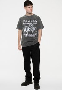 Graphic t-shirt in faded grey with black text and images. Paired with black pants and black lace-up boots. Model stands against a plain background.