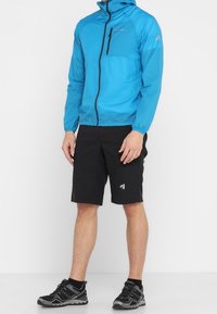 Blå letvægtsjakke med hætte, lynlåslomme foran og kontrasterende sorte shorts, parret med sorte sportssko.
