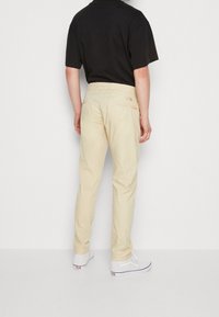 Beige Hose mit geradem Bein, aus weichem Stoff, ausgestattet mit Gesäßtaschen und einem dezenten Logodetail, kombiniert mit weißen Sneakers.