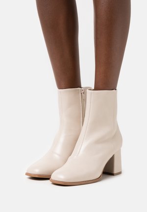 Botines de piel beige hasta el tobillo con una textura suave, cremallera lateral y tacón cuadrado, que presentan puntera redonda y un detalle de costura mínima.