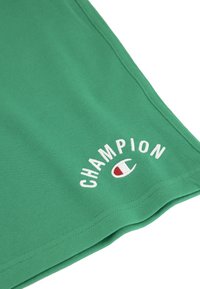 Grüne Baumwollshorts mit einem weißen, gestickten "CHAMPION"-Logo und einem kleinen roten Emblem. Weiche Textur mit genähten Kanten.
