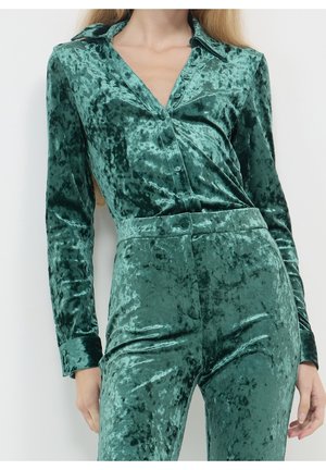 Femme portant une chemise boutonnée en velours écrasé vert brillant et un pantalon taille haute assorti sur un fond uni clair.