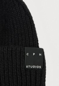 Fekete kötött sapka bordázott mintával, téglalap alakú címkével, fehér felirattal: "C P H STUDIOS."