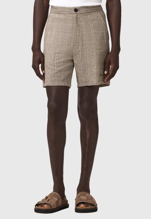Mand iført beige teksturerede shorts, brune slip-on sandaler og en hvid skjorte, stående mod en ensfarvet lys grå baggrund.