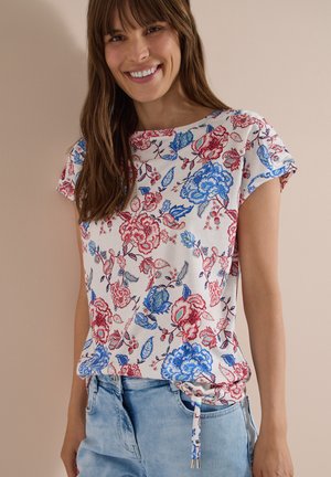 Kvinde smilende iført en hvid top med korte ærmer med rødt og blåt blomsterprint og lyseblå jeans mod en beige baggrund.