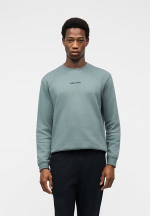 Mann trägt ein gedämpft grünes Sweatshirt mit Rundhalsausschnitt und der Aufschrift "BJÖRN BORG" sowie schwarze Hosen, steht vor einem einfarbigen hellen Hintergrund.