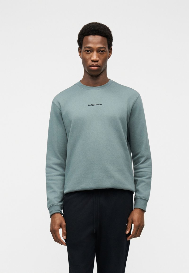 Άνδρας φοράει αχνό πράσινο φούτερ με στρογγυλή λαιμόκοψη και το κείμενο «BJÖRN BORG», καθώς και μαύρο παντελόνι, στεκόμενος μπροστά σε απλό φωτεινό φόντο.