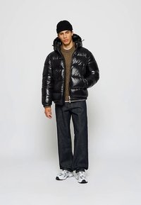 Ung mann iført svart lue, blank svart dunjakke, brun genser, mørke jeans og hvite sneakers som står mot en ensfarget bakgrunn.