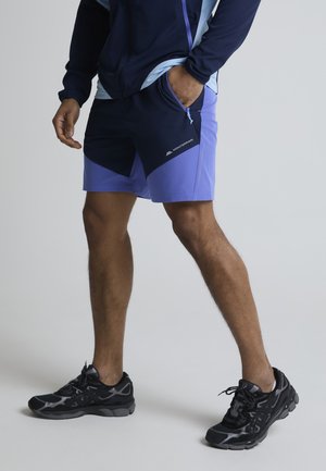 Uomo che indossa pantaloncini sportivi blu navy e viola e scarpe da corsa nere, in piedi con una mano in tasca su uno sfondo semplice.