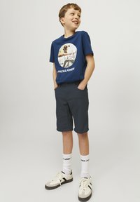 T-shirt bleu marine avec un imprimé graphique portant le texte "JACK & JONES", associé à un short gris foncé et des chaussettes blanches, des baskets crème avec des accents noirs.