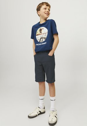T-shirt bleu marine avec un imprimé graphique portant le texte "JACK & JONES", associé à un short gris foncé et des chaussettes blanches, des baskets crème avec des accents noirs.