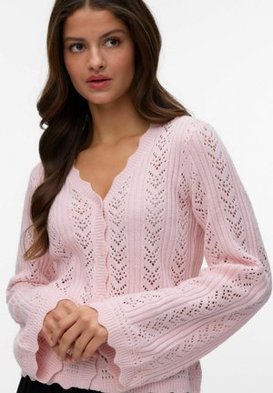 Femme aux longs cheveux bruns portant un cardigan en tricot rose clair avec des bords festonnés et un motif œillet, les mains jointes devant.