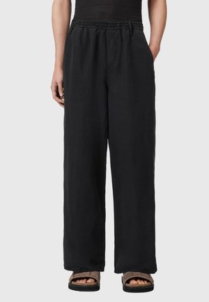 Person trägt schwarze Palazzohose mit elastischem Bund und braunen Sandalen, steht vor einem einfarbigen Hintergrund.