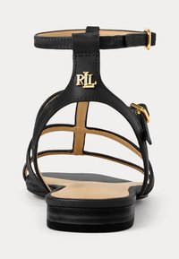 Sandal preto de pele com parte traseira aberta, faixa ajustável no tornozelo e detalhe em logo dourado. Palmilha suave e salto baixo. Design minimalista com cruzamento.