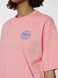 Persoon die een roze T-shirt met ronde hals draagt met een klein blauw bij-embleem en de tekst "1923 Sportswear" op de borst.
