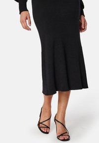 Robe en tricot noir à côtes avec une jupe évasée et un corsage ajusté, associée à des escarpins noirs à fines brides.