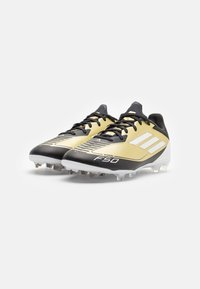 Scarpe da calcio nere e dorate con una parte superiore testurizzata, tre strisce bianche in evidenza e una suola rigida con tacchetti in plastica per una migliore trazione.