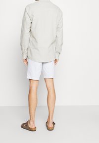 Muž stojící zády, oblečený do béžové košile s dlouhým rukávem, bílých šortek a hnědých sandálů se dvěma pásky na jednobarevném pozadí.