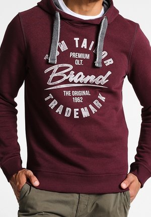 Mężczyzna w bordowej bluzie z kapturem z szarymi sznurkami i białym napisem „Tom Tailor Brand Premium Qlt. The Original 1962 Trademark.”
