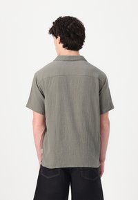 Camisa de manga corta en tejido verde oliva texturizado, con cuello con muesca y un corte relajado, con costuras visibles en la parte posterior.
