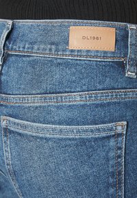 Jeans de mezclilla azul oscuro con una etiqueta de cuero que dice "DL1961" en la parte trasera, con costuras en contraste y un diseño clásico de cinco bolsillos.