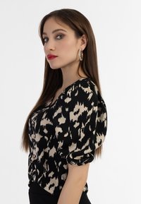 Sort og tan mønstret bluse med puffede ærmer, V-hals design, blødt stof, featuring dekorative knapper og en flydende silhuet.