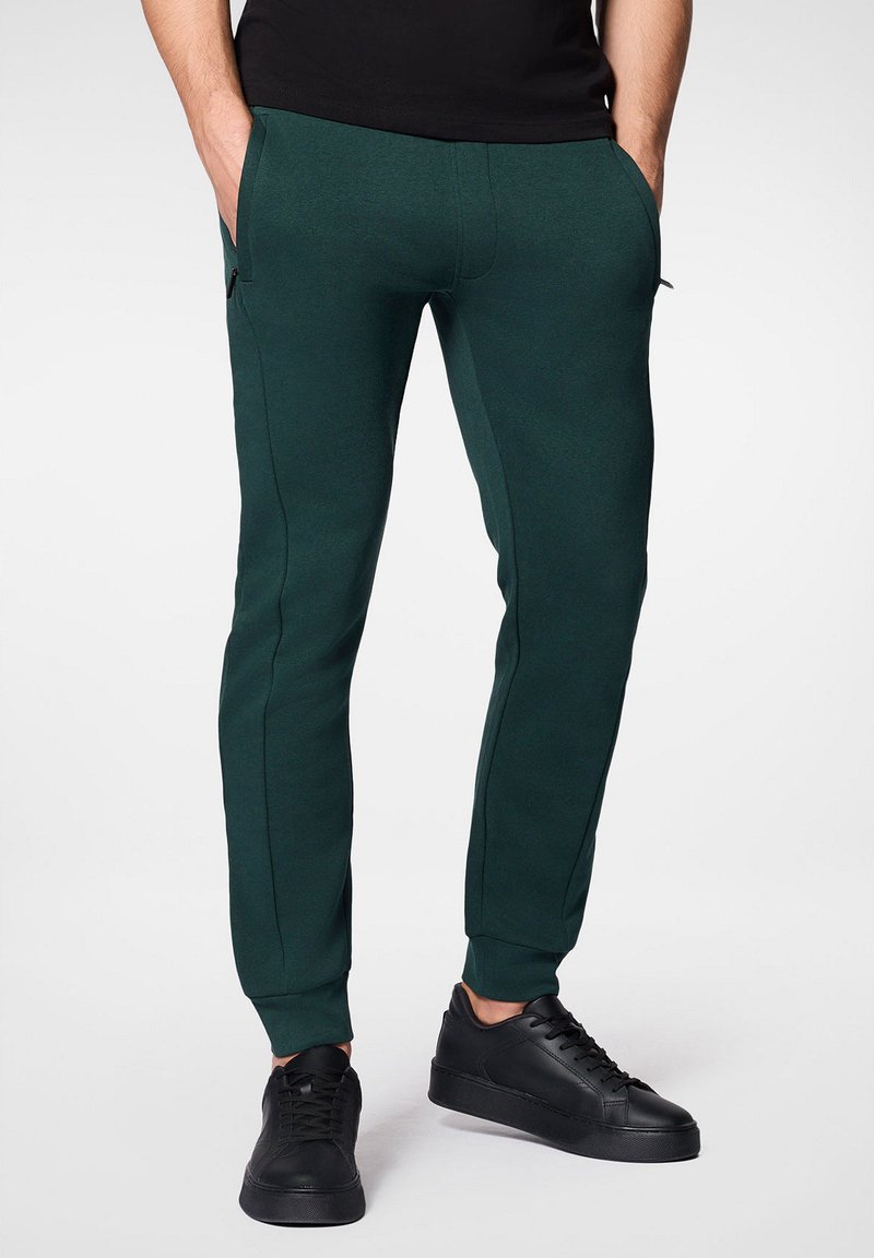 Zielone dresowe spodnie sportowe o dopasowanym kroju, wykonane z miękkiego materiału, z bocznymi kieszeniami i ściągaczami na nogawkach, w połączeniu z czarnymi trampkami.