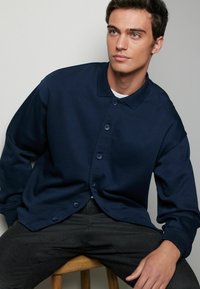 Cardigan negru închis, dintr-un amestec de bumbac, cu nasturi și guler. Croială relaxată, cu manșete ribbate și textură netedă. Purtați peste un tricou alb.