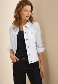 Vit denimjacka med två bröstfickor, knappstängning och uppvikta ärmar. Kombinerat med en mörk topp och mörka denimbyxor.