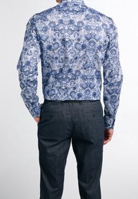 Camicia a maniche lunghe con un motivo floreale blu su sfondo bianco, abbinata a pantaloni grigio scuro. La camicia ha un colletto classico e una vestibilità sagomata.