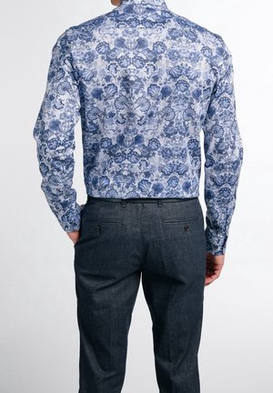 Camicia a maniche lunghe con un motivo floreale blu su sfondo bianco, abbinata a pantaloni grigio scuro. La camicia ha un colletto classico e una vestibilità sagomata.