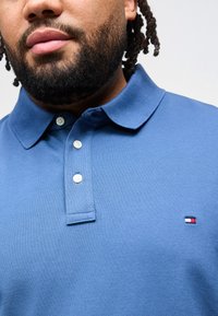 Άνδρας με γενειάδα και πλεγμένα μαλλιά, φορά μπλε πόλο μπλουζάκι με τρία λευκά κουμπιά και μικρό κόκκινο, λευκό και μπλε λογότυπο στο στήθος.