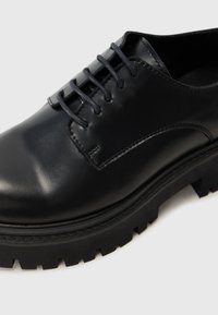 Chaussure en cuir noir à lacets avec une finition lisse, des coutures contrastantes et une semelle noire épaisse et texturée.