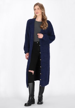 Lungo cardigan blu navy realizzato in un morbido materiale testurizzato con grandi bottoni, indossato su una maglia grigia e jeans neri strappati, abbinato a stivali neri.